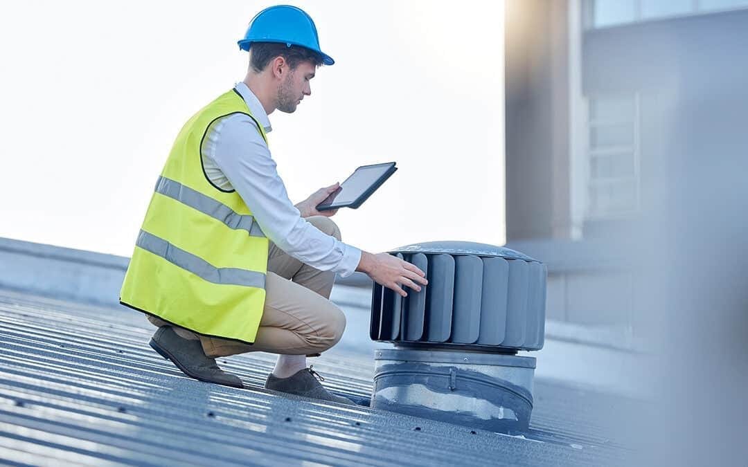 Maintenance worker in hard hat and hi-vis vest inspecting rooftop ventilation unit during a planned preventative maintenance visit